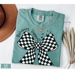 May include: Light green t-shirt with a large black and white checkered bow graphic. The words "Behavior Support" are written above the bow in a script font. A delicate gold necklace with a pendant is draped on the shirt.