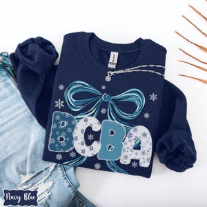May include: Navy blue sweatshirt with a large teal bow and the letters "BCBA" in a snowflake design. The sweatshirt has a crew neck and long sleeves. A silver necklace with a pendant is draped on the sweatshirt.