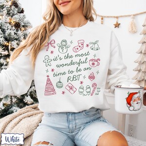 May include: White sweatshirt with a festive design. The design includes a snowman, stocking, bells, gingerbread man, and the text "it's the most wonderful time to be an RBT". A mug with a Santa Claus graphic is held in the hand.