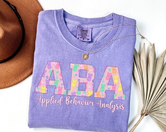 May include: Purple t-shirt with the letters ABA in pink, surrounded by colorful pastel Peeps candy. The text "Applied Behavior Analysis" is written below the letters.