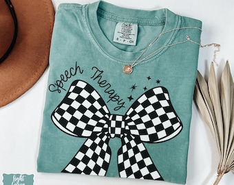 May include: A light green t-shirt with the words "Speech Therapy" above a large black and white checkered bow. A silver necklace with a small pendant is on the shirt. The shirt is folded and has a small tag.