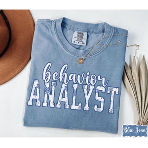May include: A blue Comfort Colors t-shirt with the words "behavior ANALYST" in white and blue script and block letters. The word "ANALYST" has snowflake accents. A gold necklace and a brown hat are also visible.