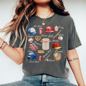 May include: A gray t-shirt with a baseball-themed design. The design includes various baseball-related items, such as a baseball bat, a baseball glove, a baseball cap, a baseball, a hot dog, a pretzel, a can of soda, and a popcorn bucket. The words "At Bat", "Walk", "Strike", "Home Run", "Inning", "Single", "Dugout", "Triple", "Strike Out", "Base", and "Double" are written around the design.