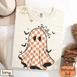 May include: Ivory-colored t-shirt with a distressed look. The shirt features a ghost design with an orange and white checkered pattern, the letters "BCBA", and several black bats. A gold necklace and a hat are visible.