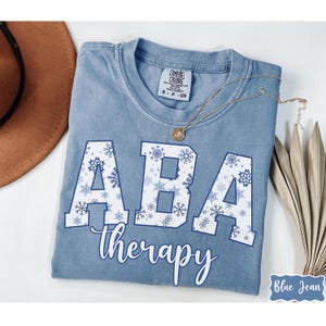 May include: A blue Comfort Colors t-shirt with the words "ABA therapy" in white and snowflake accents. The shirt has a crew neck and short sleeves. A gold necklace is draped across the shirt.