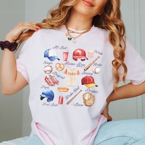 May include: A white t-shirt with a baseball-themed design. The design features a heart shape with various baseball-related items, including a baseball, bat, glove, hat, and a baseball jersey. The words "At Bat", "Walk", "Strike", "Single", "Home Run", "Dugout", "Triple", "Inning", "Strike Out", "Double", "Base", and "Foul Ball" are written around the heart.
