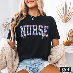 Puede incluir: Una camiseta negra con la palabra "NURSE" impresa en letras blancas. Las letras est&aacute;n formadas por gr&aacute;ficos de paletas de hielo rojas, blancas y azules. La camiseta la lleva puesta una persona con el pelo rubio y se combina con unos pantalones cortos de mezclilla.