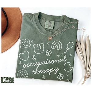 May include: A moss green t-shirt with the words "occupational therapy" in white script, surrounded by white graphic icons. A gold necklace with a pendant rests on the shirt. A brown hat and dried plant are in the background.