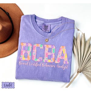 May include: A purple t-shirt with the text "BCBA Board Certified Behavior Analyst" printed on the front. The letters are decorated with a pattern of colorful Peeps marshmallow candies.