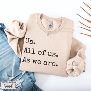 May include: A beige sweatshirt with the text "Us. All of us. As we are." printed in black.