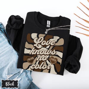 May include: Black sweatshirt with a retro-style graphic that says "Love knows no color" with hearts that say "Proud" and "Equal".