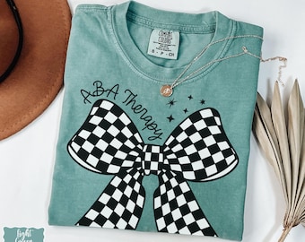 May include: Light green t-shirt with the text "ABA Therapy" above a large black and white checkered bow. A gold necklace with a pendant is draped across the shirt. The shirt has a Comfort Colors tag.