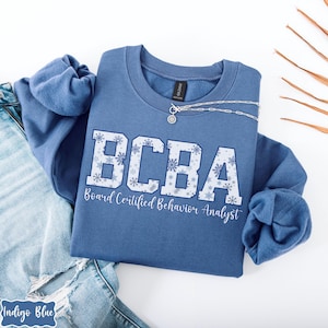 May include: A blue crewneck sweatshirt with the text "BCBA" in white block letters with snowflake accents. Below the letters is the text "Board Certified Behavior Analyst" in a script font. A silver necklace with a pendant is on the sweatshirt.