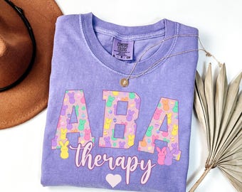 May include: Purple t-shirt with the text "ABA therapy" in white with a pink heart. The letters are filled with a pattern of pastel colored Peeps candy.