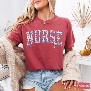 Puede incluir: Camiseta roja con la palabra "NURSE" en letras blancas con un patr&oacute;n de polos de helado rojo, blanco y azul. La camiseta la lleva puesta una persona con el pelo rubio y se combina con unos pantalones cortos de mezclilla.