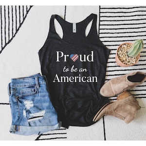 Puede incluir: Camiseta de tirantes negra con un gr&aacute;fico blanco que dice "Proud" con un coraz&oacute;n de bandera estadounidense y "to be an American".