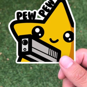May include: A yellow star-shaped sticker with a cartoon face and the word "PEW" in black and white. The star is holding a grey and black toy gun. The sticker is held by a hand against a green background.