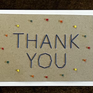 May include: A handmade card with a brown background and a white border. The words "THANK YOU" are stitched in blue thread. The card is decorated with small colorful dots.