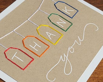 May include: A handmade thank you card with a kraft paper background and a white border. The words "THANK YOU" are embroidered on colourful tags. The word "you" is written in cursive with white thread.
