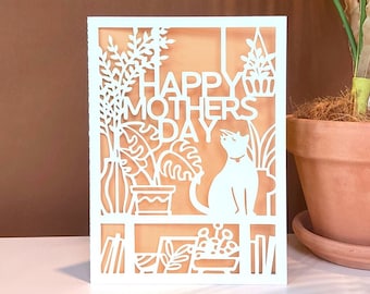 May include: White card with a cut-out design of a cat sitting on a shelf with plants and books. The card says "Happy Mother's Day" in a white font.