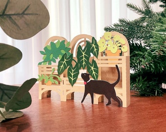 May include: A papercraft model of a black cat with a red collar walking past a miniature wooden shelf with potted plants. The shelf is decorated with a green, brown, and yellow color scheme.