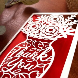 May include: A red card with a white cutout design of a vase with flowers and the words "Thank You".