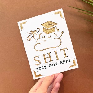 Peut inclure: Une carte blanche avec des accents de paillettes dor&eacute;es repr&eacute;sente un nuage de dessin anim&eacute; avec un bonnet de graduation et le texte "SHIT JUST GOT REAL".