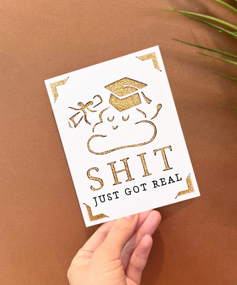 Peut inclure: Une carte blanche avec des accents de paillettes dor&eacute;es repr&eacute;sente un nuage de dessin anim&eacute; avec un bonnet de graduation et le texte "SHIT JUST GOT REAL".