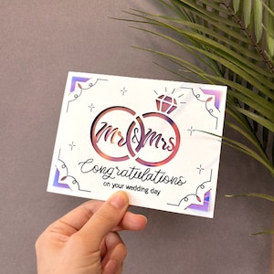 May include: A white card with a holographic finish features a cut-out design of two interlocking rings with the words "Mr & Mrs" inside. The card also includes the text "Congratulations on your wedding day".