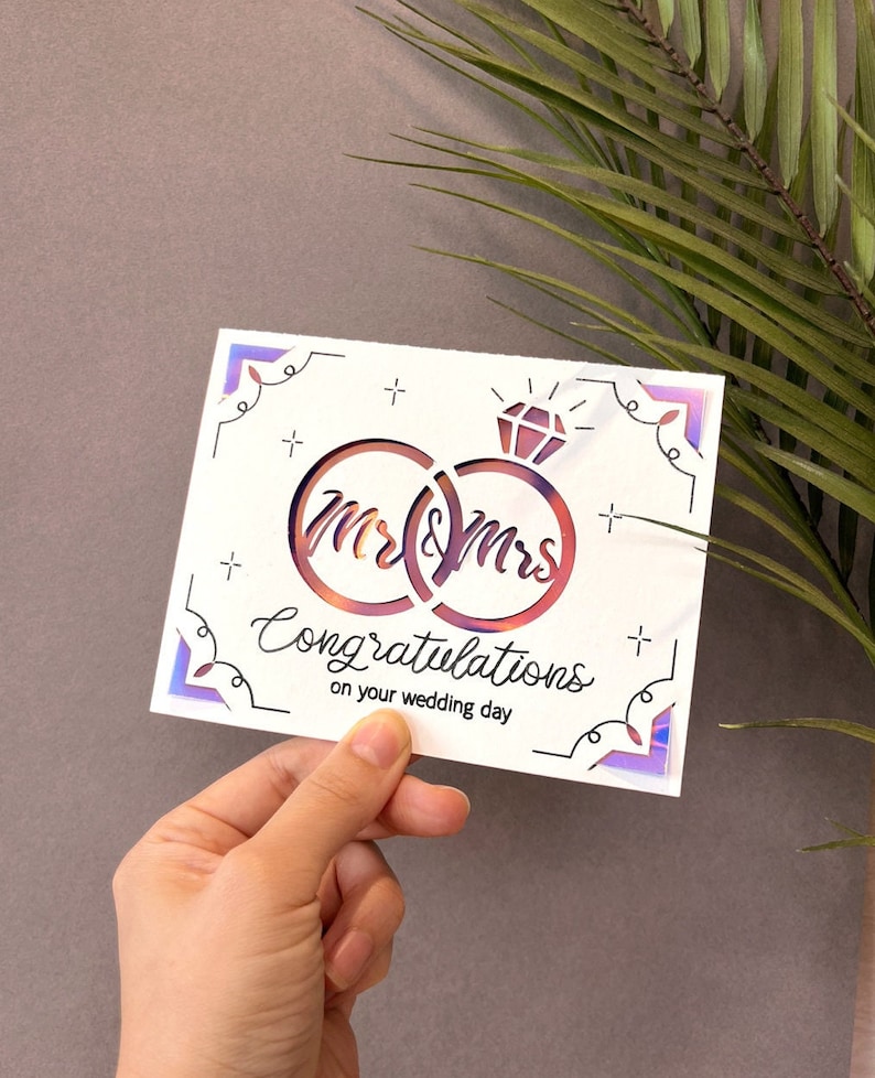 May include: A white card with a holographic finish features a cut-out design of two interlocking rings with the words "Mr & Mrs" inside. The card also includes the text "Congratulations on your wedding day".