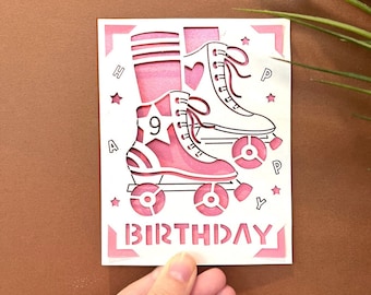 May include: Pink birthday card with a cut-out design of roller skates and the words "Happy Birthday".