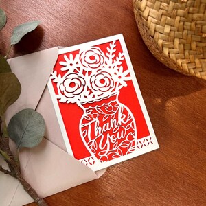 May include: A red card with a white cutout design of a vase with flowers and the words "Thank You".