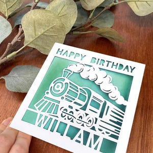 May include: A white card with a green background and a cut-out design of a train engine. The card says "Happy Birthday William" in white lettering.