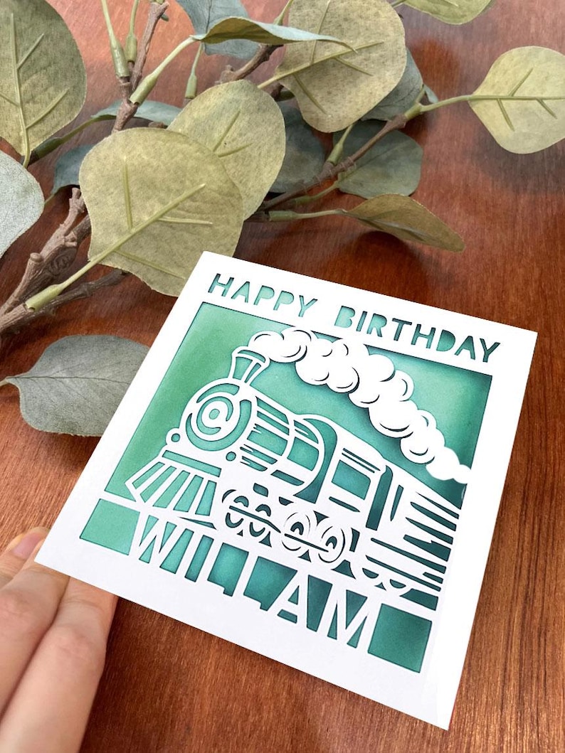 May include: A white card with a green background and a cut-out design of a train engine. The card says "Happy Birthday William" in white lettering.