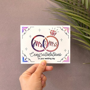 May include: A white card with a pink and blue iridescent border. The card features a graphic of two interlocking rings with the words "Mr & Mrs" inside. The text "Congratulations on your wedding day" is written below the rings.