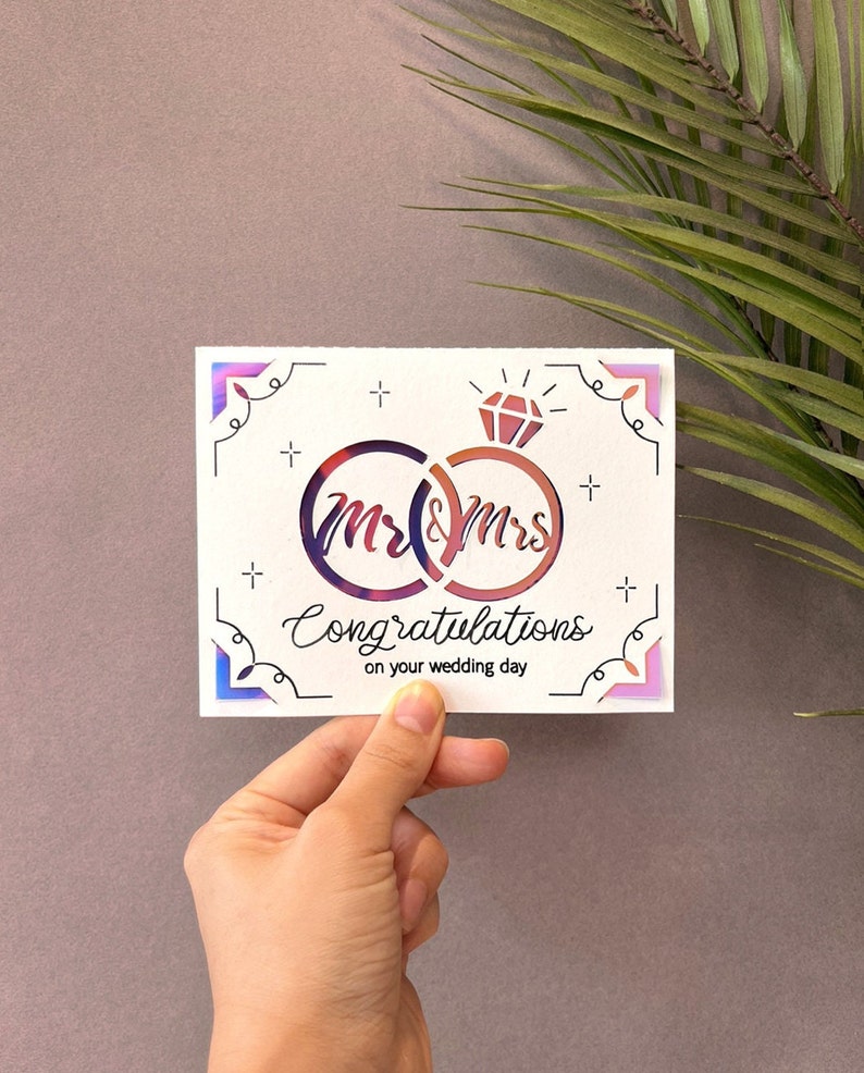 May include: A white card with a pink and blue iridescent border. The card features a graphic of two interlocking rings with the words "Mr & Mrs" inside. The text "Congratulations on your wedding day" is written below the rings.