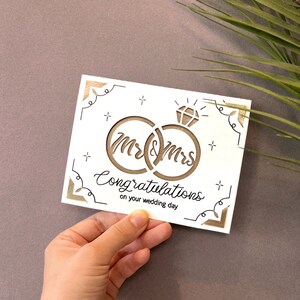 May include: White card with a brown cutout design of two interlocking rings with a diamond on top. The rings have the words "Mr & Mrs" written inside. The card also has the text "Congratulations on your wedding day".