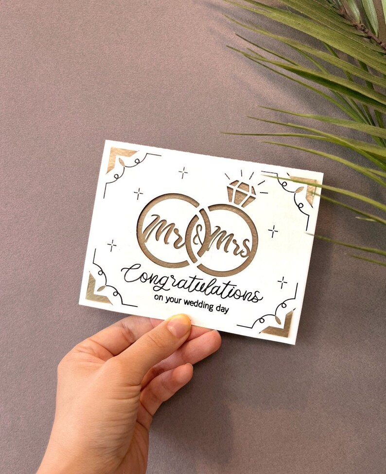 May include: White card with a brown cutout design of two interlocking rings with a diamond on top. The rings have the words "Mr & Mrs" written inside. The card also has the text "Congratulations on your wedding day".