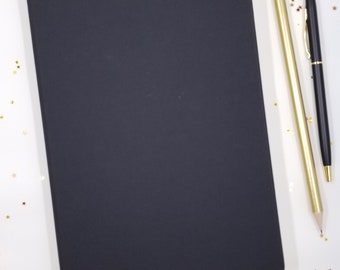 May include: A black notebook with a black elastic band closure, a gold pencil with a pink eraser, and a black pen on a white surface with gold glitter stars scattered around.