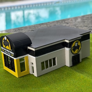 May include: A 3D printed scale model of a Buffalo Wild Wings Grill & Bar restaurant building. The structure features a black roof, light gray walls, a yellow entrance section, and black signage with the Buffalo Wild Wings logo and text.