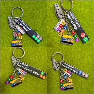 May include: A collection of four metal keychains featuring New York City subway themes. Each keychain includes a silver train charm, a rectangular station sign with text, a yellow MetroCard charm, and small circular coloured line indicator charms.