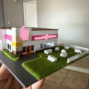 May include: A scale model of a commercial building featuring white walls, orange door frames, and a pink horizontal stripe. The model includes a green lawn area with several small white picnic tables and a white picket fence surrounding the perimeter.