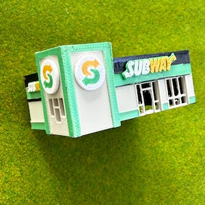 May include: A miniature 3D printed model of a Subway restaurant building. The structure is white and light green with a black roof trim. It features the Subway logo on the front and side, with the word Subway in yellow letters on a black background.