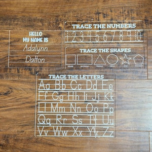 Op de afbeelding: Transparante acryl traceerborden voor educatieve doeleinden. Inclusief borden voor het traceren van cijfers, vormen, letters en naam oefening. De borden bevatten de tekst "TRACE THE NUMBERS", "TRACE THE SHAPES", "TRACE THE LETTERS", en "HELLO MY NAME IS".