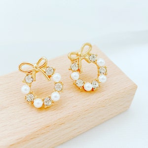 May include: A pair of gold tone earrings with a wreath design. The wreath is adorned with white pearls and clear rhinestones. The earrings have a bow detail at the top.
