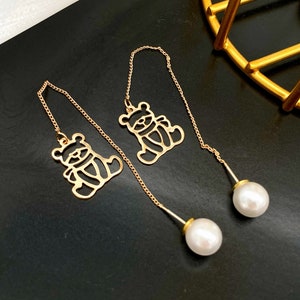 May include: Gold-colored drop earrings featuring a teddy bear silhouette. The earrings have a delicate chain design, with a large white pearl at the end of each earring. The earrings are displayed on a black surface.