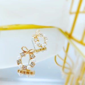 May include: A pair of gold-tone earrings with a pearl and crystal wreath design. The earrings feature a small bow at the top of the wreath.