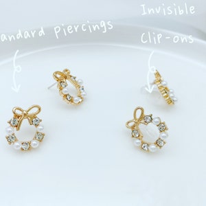 May include: Three pairs of gold-tone earrings with a pearl and rhinestone wreath design. The earrings are shown in three styles: standard piercings, clip-ons, and invisible clip-ons.