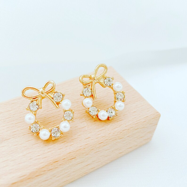 May include: A pair of gold tone earrings with a wreath design. The wreath is adorned with white pearls and clear rhinestones. The earrings have a bow detail at the top.
