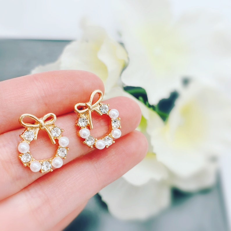 May include: A pair of gold-tone earrings with a wreath design. Each earring features a small bow and is adorned with white pearl-like stones.